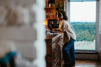 A woman wearing pink headphones leans against a kitchen counter while working on a laptop near a large window, representing parenting tips for a child with ADHD.
