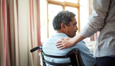 A caregiver places a supportive hand on the shoulder of an elderly man sitting in a wheelchair.