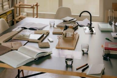 A large table is covered with various architectural tools and materials, including a house model, fabric swatches, open notebooks, and glasses of water.