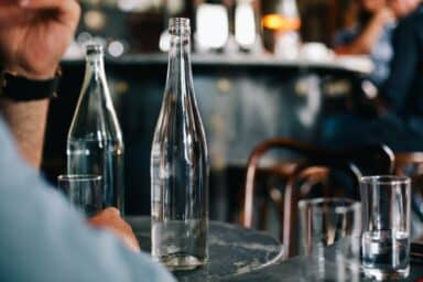 Empty clear glass bottle on a dark bistro table in a bar setting, representing the isolation of alcoholism and alcohol abuse.