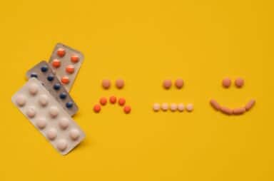 Various blister packs and loose pills arranged on a bright yellow surface to form a sad face, a neutral face, and a smiling face, representing antidepressants.