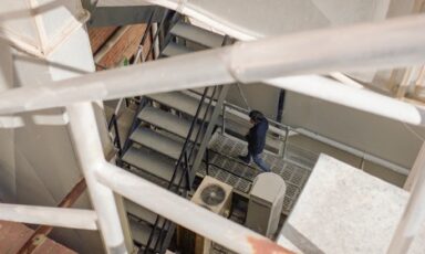 A person in a dark jacket and jeans walks down a set of metal industrial stairs, viewed from a high angle through white railings.