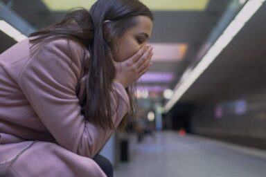 A young woman in a pink coat sits on a subway platform, covering her face with her hands in distress.