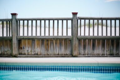 A weathered wooden fence stands between a bright blue swimming pool and white sand dunes under a clear sky.