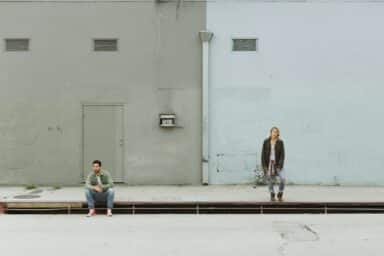 Two individuals seated on the sidewalk in front of a building, engaged in conversation and enjoying their surroundings.