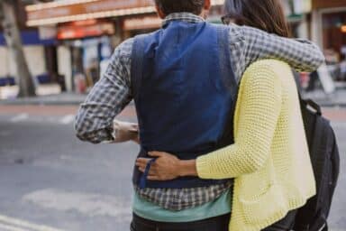 A woman in a bright yellow sweater walks with her arm around a man in a blue vest on a city street.