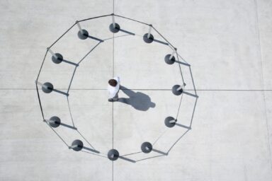 An overhead view shows a single person standing in the center of a circle formed by stanchions and rope on a large, open concrete surface.