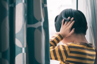 A child in a striped shirt covers their ears with their hands while looking out a window, illustrating sensory sensitivity in autism.