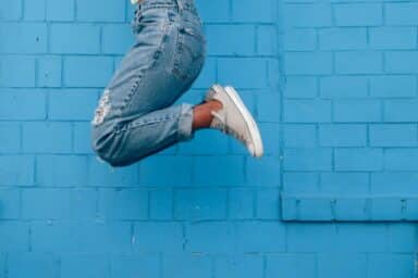 A person wearing denim jeans and white sneakers jumps high in the air against a bright blue brick wall, representing the benefits of play for adults.