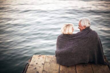 An older couple wrapped in a dark blanket sit on a wooden dock overlooking the water, illustrating intimacy and better sex as you age.