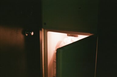 A refrigerator door stands slightly ajar in a dark room, casting a warm glow from the interior, representing binge eating disorder.