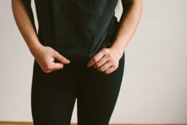 A person in a black t-shirt and black pants stands against a plain, light-colored wall, their hands gripping and pulling at the hem of their shirt near their waist.