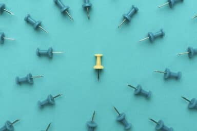 An illustration features a single yellow pushpin standing upright in the center of a blue surface, surrounded by several blue pushpins that are tipped over and pointing toward it.