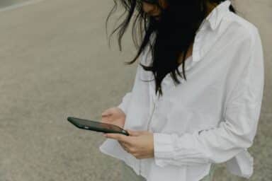 A woman in a white button-down shirt is looking down at a smartphone she is holding in both hands.