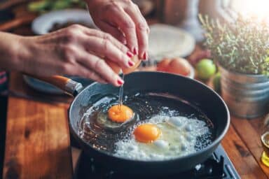 A person with red nail polish cracks an egg into a black frying pan on a wooden counter, representing choosing healthy protein.