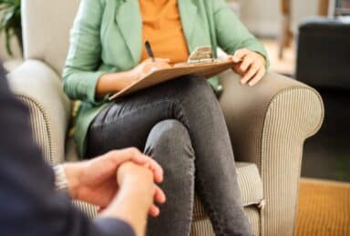 A therapist in a green blazer sits in an armchair across from a client, holding a clipboard and pen to take notes during a session.