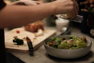 A person's hands grating fresh Parmesan cheese over a garden salad with tomatoes in a ceramic bowl.