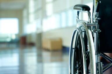 A manual wheelchair sits in the foreground of a blurred, brightly lit hospital or clinic hallway.