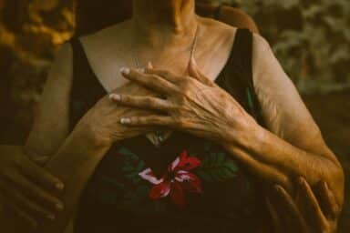 An older adult wearing a floral top rests their hands over their heart while being embraced from behind.