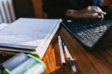 Close-up of a person’s hands typing on a laptop next to a notepad with handwritten notes and two pens on a wooden desk.