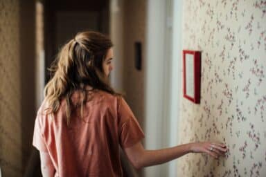 A woman in a peach shirt stands in a hallway, reaching out to touch floral-patterned wallpaper.