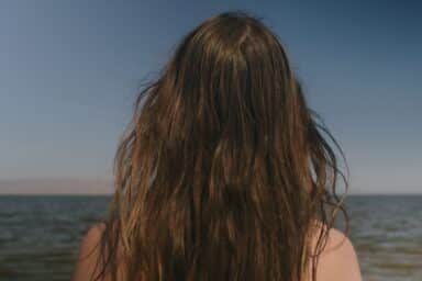 Back view of a person with long brown hair looking out at a calm sea under a clear blue sky.