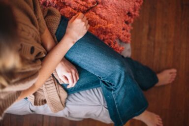A woman in a tan cardigan and blue jeans sits on a grey bed with her legs crossed, representing the experience of depression in women.