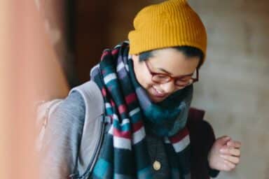 A person wearing a yellow beanie, glasses, and a multi-colored striped scarf smiles while looking down, representing depression treatment.