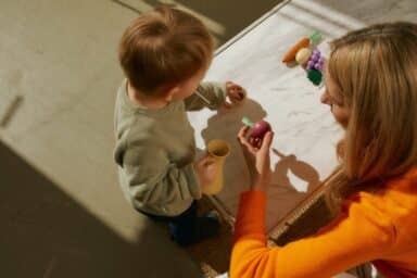 High-angle shot of a young child in a green sweater playing with a woman in an orange top at a white table, representing early autism screening.