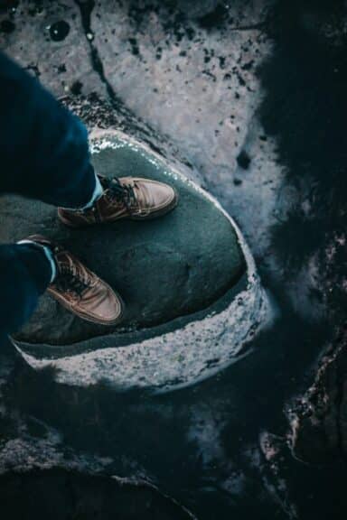 A person’s feet in brown boots stand on the edge of a large, dark rock surrounded by water, representing the precarious nature of drug abuse and addiction.