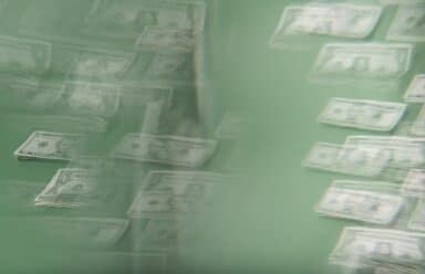 An abstract, blurred image shows several stacks of U.S. dollar bills against a green background, with a hazy, double-exposure effect creating a sense of movement or confusion.