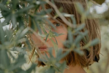 A close-up image shows a person's face partially obscured by the green leaves and branches of a tree, with one blue eye looking directly through the foliage.