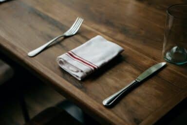 A simple place setting with a fork, a folded white napkin with red stripes, a knife, and a glass on a wooden table, representing emotional eating.