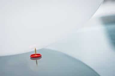 A white balloon is shown hovering dangerously close to the sharp point of a red pushpin resting on a reflective surface.