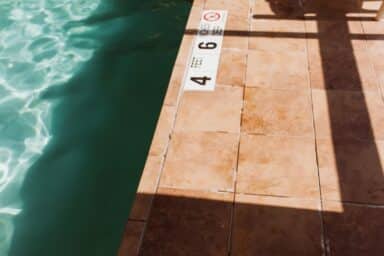 Edge of a swimming pool with tan tiles and turquoise water, where depth markers read "4 FEET 6 INCHES DEEP."