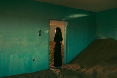 Person in a long black dress stands in a doorway looking into a room filled with sand, while more sand is piled up in the teal-walled room where they are standing.