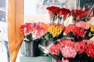 Bunches of red, yellow, and pink roses are wrapped in plastic and displayed in a flower shop window next to a bamboo blind.