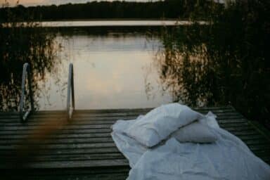 White pillows and a blanket are spread out on a wooden dock overlooking a calm lake at twilight.