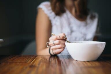 A woman in a white lace top sits at a wooden table, holding a spoon over a white bowl, representing healthy eating for women.