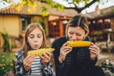 A woman and a young girl sit outdoors together, each happily eating a large cob of corn, representing healthy food for kids.