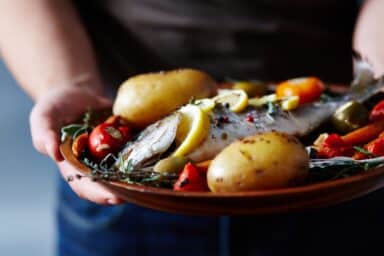 A person holding a wooden plate filled with a cooked fish topped with lemon slices, potatoes, olives, and herbs, representing a heart-healthy diet.