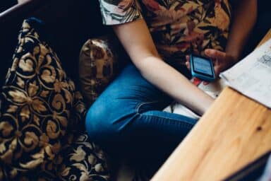 A person in a floral shirt and jeans sits on a sofa, looking at a smartphone next to a wooden table.