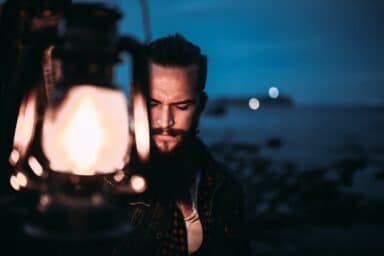 A man with a beard looks down pensively while holding a glowing lantern at night near a dark shoreline.