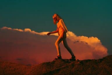 A woman in a white jumpsuit walks across a rocky landscape under a dramatic orange and blue sunset, representing helping someone with an eating disorder.