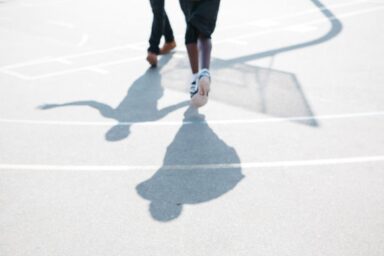 Low-angle view of two people walking on an outdoor basketball court, focusing on their legs and long shadows.