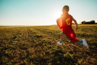 A young child wearing a red superhero cape runs through a sun-drenched grassy field toward the horizon, representing helping your child with autism thrive.