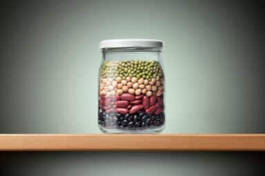 A glass jar filled with layers of black beans, kidney beans, chickpeas, and green lentils sits on a wooden shelf, representing high-fiber foods.
