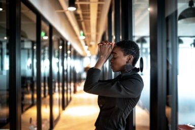 A woman with short hair and large earrings leans against an office wall with eyes closed and hands clasped to her forehead in a moment of stress or deep focus.