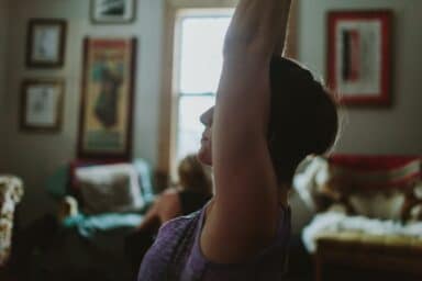 A woman in a purple tank top reaching her arms overhead while practicing yoga in a dimly lit living room.