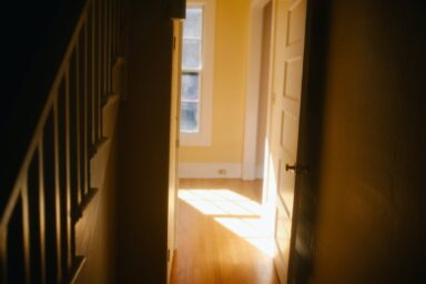 Warm sunlight spills across a hardwood hallway floor from an open doorway in a dimly lit home interior near a staircase.